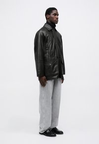 Veste en cuir noir avec col, poches avant et fermeture par bouton, associée à un jean gris à jambes larges et des mocassins noirs.