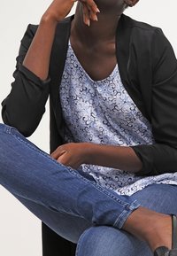 Černý blazer přes modrý vzorovaný top, modré džíny a černé ploché boty. Top má květinový vzor se spirálami.