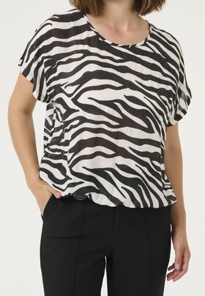 Frau trägt eine lockere, schwarz-weiße, zebra-gestreifte Kurzarmbluse und schwarze Hose, steht mit einer Hand in der Tasche.