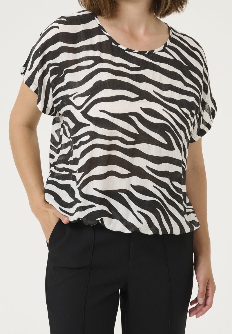 Femme portant un chemisier ample à manches courtes noir et blanc à rayures zèbre et un pantalon noir, debout avec une main dans la poche.