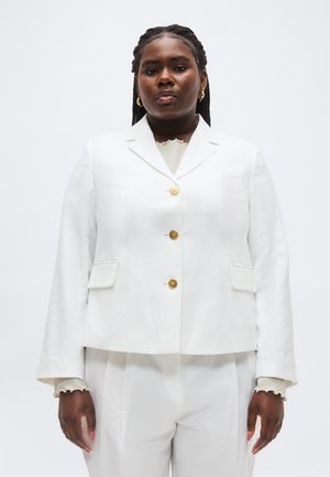 Vrouw met gevlochten haar, gouden oorringen en een witte blazer met drie knopen, gecombineerd met een bijpassende witte broek.