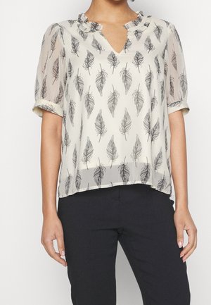 Femme portant un chemisier blanc transparent avec des imprimés plumes noirs, col à volants et manches retroussées, associé à un pantalon noir.