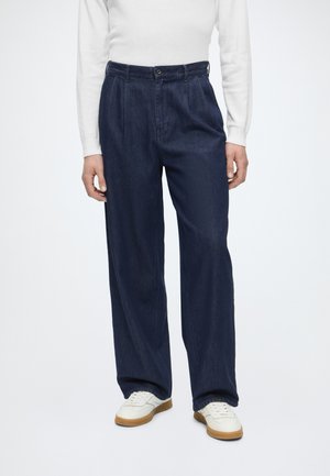 Personne portant un jean large bleu foncé, un haut blanc à manches longues et des baskets blanches avec semelles en gomme, debout devant un fond uni.