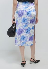 Femme portant une jupe midi florale violette et bleue avec une fente latérale, des talons slingback noirs, et tenant un sac à main noir.