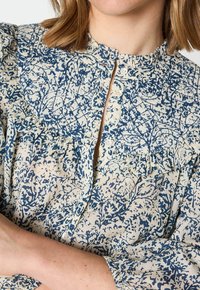 Blus med blommönster i kräm och marinblått, med volangdetaljer, en knappstav och hög hals. Texturen verkar lätt.