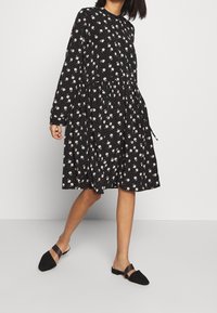 Svart långärmad klänning med vitt blommönster, skrynklig midja och utställd kjol. Matchad med svarta slip-on-mules med brun accentrem.