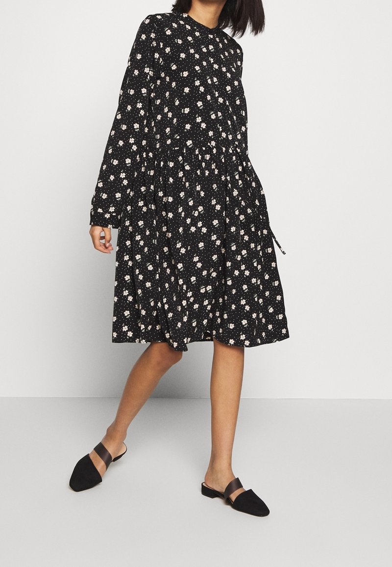 Svart långärmad klänning med vitt blommönster, skrynklig midja och utställd kjol. Matchad med svarta slip-on-mules med brun accentrem.