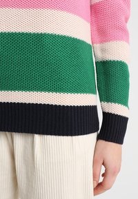 Pull en tricot à rayures horizontales rose, verte, crème et bleu marine ; tissu texturé ; poignets et ourlet côtelés ; porté avec un pantalon en velours côtelé crème.