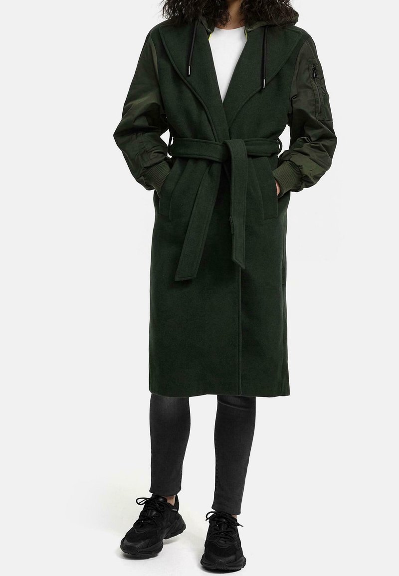 Person iført en mørkegrøn bæltefrakke med stofkrop og nylon bomber-lignende ærmer, sorte leggins og sorte sneakers.