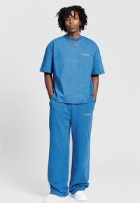 Sininen puuvillapaita lyhyillä hihoilla ja löysät siniset henkselit, joissa on huomaamattomia logotietoja, yhdistettynä valkoisiin lenkkikenkiin.