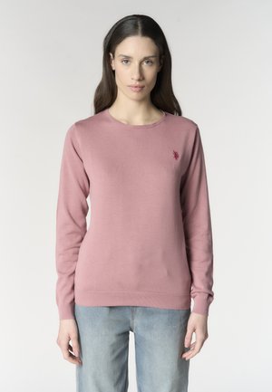 Donna con capelli castani lunghi indossa un maglione rosa a maniche lunghe e jeans azzurri, in piedi davanti a uno sfondo chiaro e semplice.