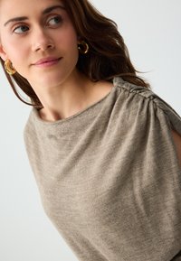 Jeune femme aux cheveux bruns portant des boucles d'oreilles créoles dorées et un haut beige en tricot avec un détail froncé à l'épaule, regardant légèrement sur le côté.