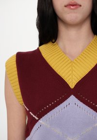 Femme portant un gilet en tricot sans manches avec un col jaune et un bord aux emmanchures, partie supérieure bordeaux et motif losanges lavande.