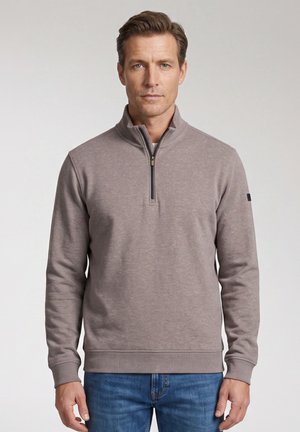 Man die een lange-mouw grijze pullover met een kwart rits en een blauwe spijkerbroek draagt, staand voor een effen lichte achtergrond.