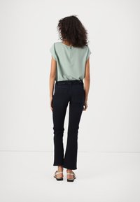 Light green short-sleeve top with a back zipper, paired with dark blue flared jeans. The model wears tan flat sandals with a decorative strap.