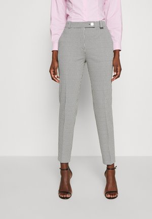 Personne portant un pantalon slim à carreaux noir et blanc, une chemise rose à manches longues et des talons ouverts noirs, debout devant un fond uni.