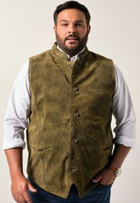 Chaleco de ante verde oliva con cuello alto y costuras decorativas. Cuenta con cinco botones y dos bolsillos frontales. Lleva puesto sobre una camisa blanca.