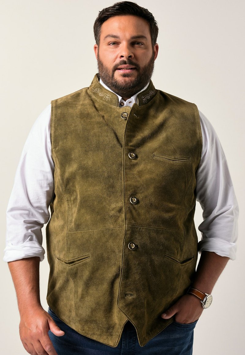 Chaleco de ante verde oliva con cuello alto y costuras decorativas. Cuenta con cinco botones y dos bolsillos frontales. Lleva puesto sobre una camisa blanca.