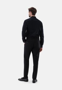 Schwarzes Langarmshirt und schwarze Hose mit glattem Stofftextur und Knopfakzenten. Die Schuhe sind schwarz und poliert.