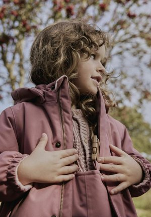 Bērns ar cirtainiem matiem, kas piesprauž mauve krāsas lietusmēteli, ģērbies adītā džemperī, brīvā dabā ar izplūdušiem kokiem fonā.