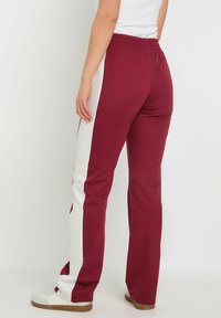 Pantalon de survêtement bordeaux avec des rayures blanches sur les côtés, taille élastique et coupe ample. Fabriqué en tissu léger et doux au toucher.