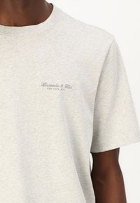 Tricou scurt cu mânecă gri, din bumbac. Dispune de un guler rotund și textul logo brodat pe piept în gri închis.