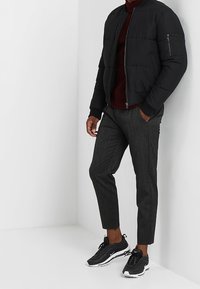 Giacca bomber imbottita nera, dolcevita bordeaux, pantaloni grigi strutturati e sneaker sportive nere con dettagli bianchi. Posizione eretta, sfondo neutro.