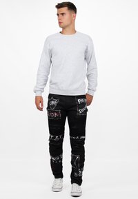 Felpa grigia e jeans neri con toppe artistiche e dettagli argentati. I jeans hanno una vestibilità slim con dettagli testurizzati lungo le gambe.