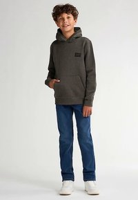 Šedá mikina s kapucí, přední kapsou a textilní štítkem, ve spojení s modrými džínami a bílými teniskami, předvedena na mladém modelu.