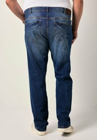 Jeans de denim azul oscuro con un corte estándar, dos bolsillos traseros y un lavado desgastado con costuras en contraste. Zapatillas blancas visibles en la parte inferior.