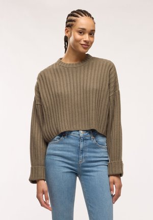 Giovane donna con i capelli intrecciati che indossa un maglione marrone a coste ampio e jeans blu a vita alta su uno sfondo chiaro uniforme.