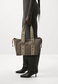 Bruine leren tote bag met crèmekleurige hengsels en logo-tekst. Het heeft een gestructureerde vorm en zijkant ritsafsluitingen, wordt met één hand gedragen.