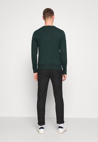 Homme debout, de dos, portant un pull vert foncé, un pantalon à carreaux noir, des baskets blanches et des chaussettes blanches, sur fond blanc uni.