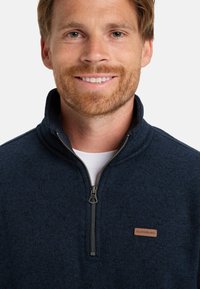 Navy zip-up fleece with a textured knit, high collar, and a small brown logo patch on the chest. Features a silver zipper detail.