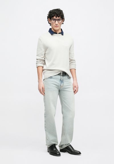 Pull-over gris clair avec col bleu marine, associé à un jean bleu clair et des chaussures habillées noires. Tricot texturé notable et détails de ceinture.