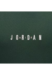 Tela verde con "JORDAN" bordado en blanco y rosa claro. La textura es suave, con un ligero estiramiento. El diseño presenta letras mayúsculas.