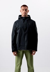 Veste noire imperméable avec capuche, dotée de poches zippées, au design minimaliste et d'un logo sur la poitrine, associée à un pantalon vert.