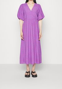 Mujer vistiendo un vestido morado hasta el tobillo con mangas abullonadas y escote en V, combinado con sandalias de plataforma negras sobre un fondo liso.