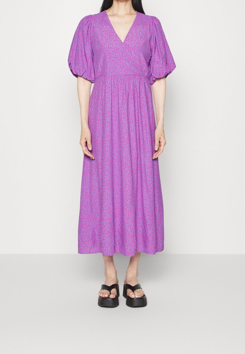 Mujer vistiendo un vestido morado hasta el tobillo con mangas abullonadas y escote en V, combinado con sandalias de plataforma negras sobre un fondo liso.