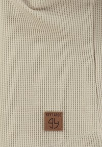 Beige ribbel gebreide stof met een klein bruin vierkant label met de tekst "KEY LARGO" en een minimalistisch logo onder de tekst.