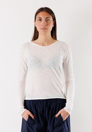 Donna con capelli lunghi e scuri in piedi rivolta in avanti, indossa una maglia bianca trasparente a maniche lunghe e pantaloni blu scuro, con le mani nelle tasche.