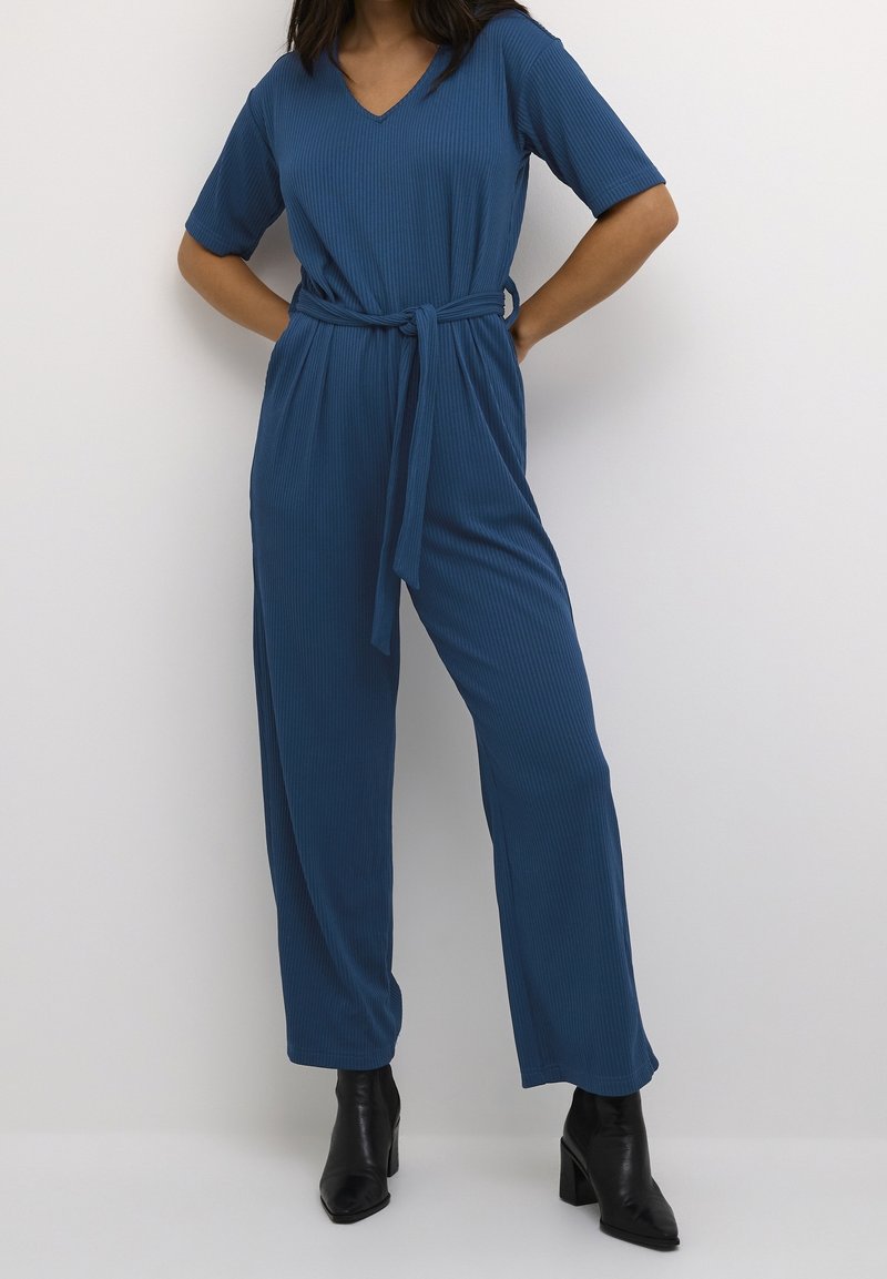Mono azul de canalé con mangas cortas, escote en V, cintura ceñida con cinturón y piernas anchas, combinado con botas de tobillo negras de tacón.