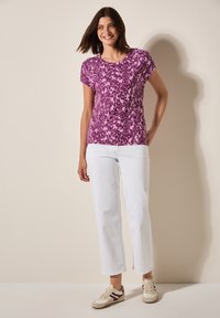 Top morado de manga corta con un patrón abstracto texturizado, combinado con pantalones blancos rectos y zapatillas beige con acentos burdeos.