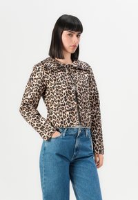 Kvinna med axellångt mörkt hår som bär en leopardmönstrad jacka och blå jeans, stående med ena handen i fickan mot vit bakgrund.