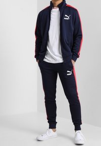 Marineblauw trainingspak met rode zijkanten, bestaande uit een ritsjack en bijpassende broek, beide met witte Puma-logo's en elastische manchetten.