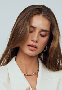 Silverpärlspets och hoops örhängen bärs med en vit blazer. Håret är långt och vågigt, med mjuka vågor som ramar in ansiktet.