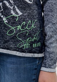 Langärmliger, grauer Pullover mit einem Farbverlauf-Textur. Verfügt über einen grünen Aufdruck mit der Aufschrift "Soccx Spirit EST. BRAND" sowie Rippenbündchen und -saum.