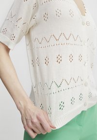 Weiße gestrickte Strickjacke mit Durchbruchs-Mustern, kurzen Ärmeln und Knopfverschluss. Der Saum hat eine leichte Kurve und strukturierte Details.