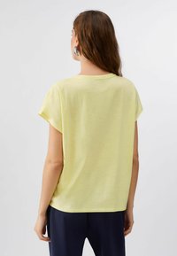 Femme aux cheveux longs bruns portant un haut à manches courtes jaune clair et un pantalon foncé, debout de dos sur un fond uni.