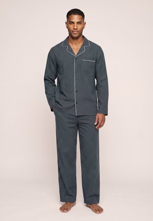 Hombre de pie descalzo vistiendo pijamas oscuros a cuadros de manga larga con botones, con ribete blanco y pantalones a juego sobre fondo claro.